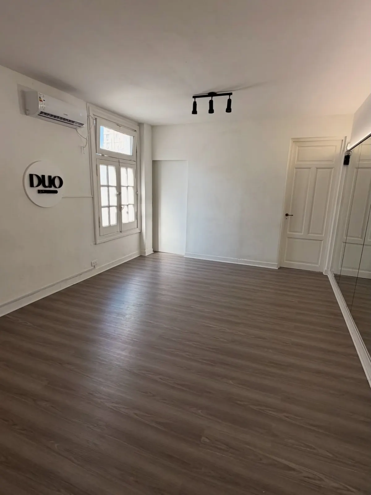 Sala vacía con piso de madera, aire acondicionado, ventana y el logo de Duo Estudio en la pared